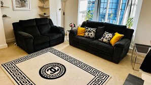 Sala de estar con dos sofás oscuros, un tapete estampado y un ventanal con vistas a edificios.