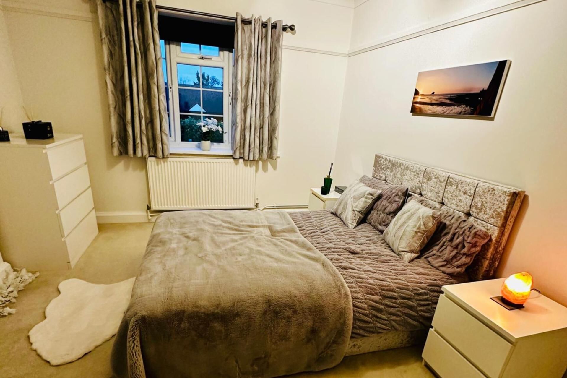 A bedroom with a bed, a nightstand, a window with curtains, a radiator, a wall-mounted picture, and a bedside lamp.
