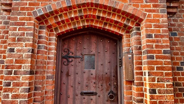 Una puerta de madera con manija metálica y un pequeño ventanal, incrustada en un muro de ladrillo con un arco decorativo arriba.