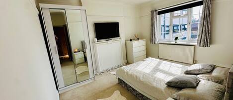 Un dormitorio con cama, mesita de noche, televisor, armario y ventana con cortinas.