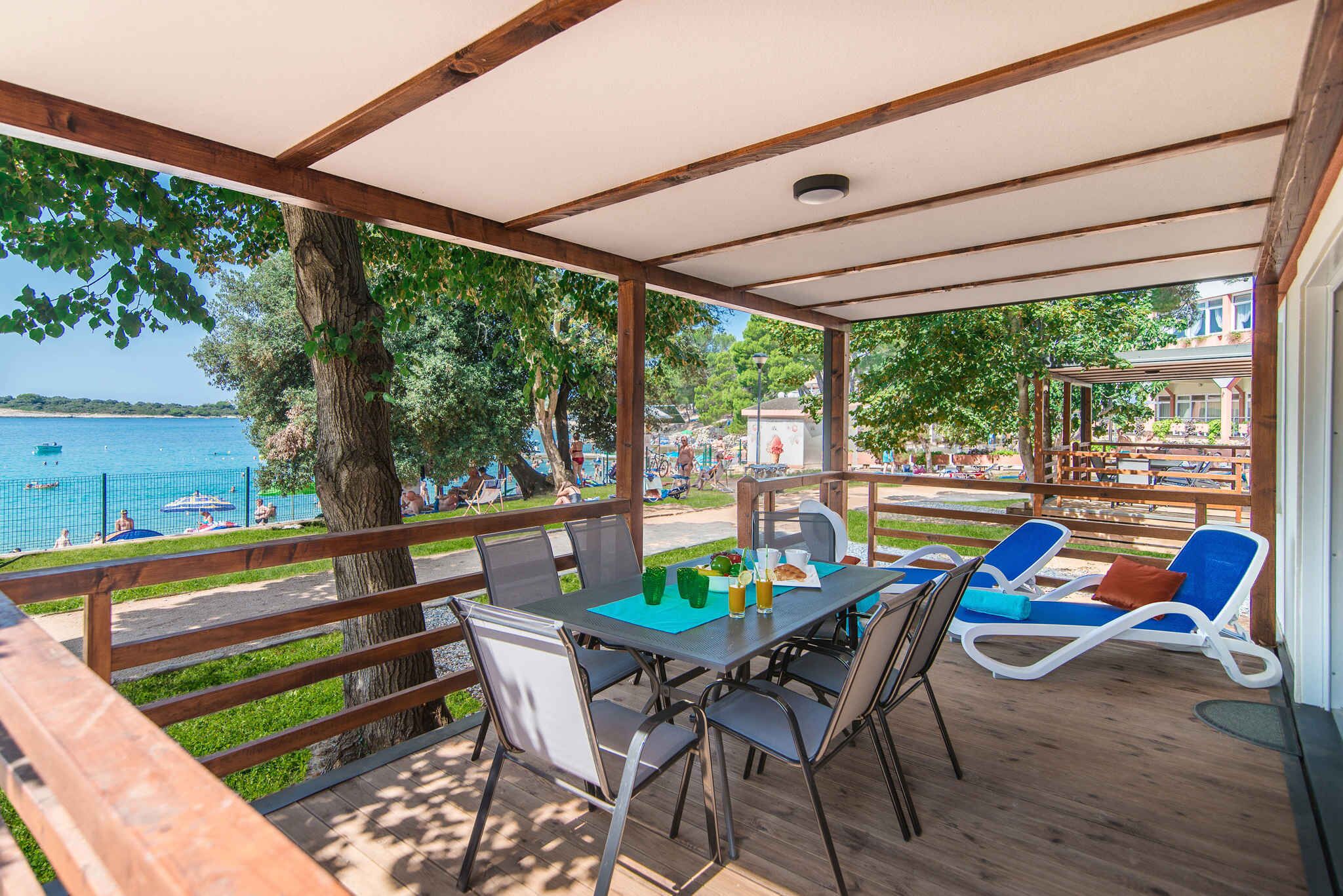 A covered outdoor seating area with a table, chairs, and lounge chairs overlooking a body of water.