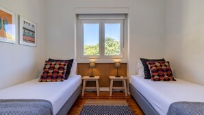 Una habitación pequeña y bien iluminada con dos camas individuales, una ventana con vistas a árboles y una mesita entre las camas.