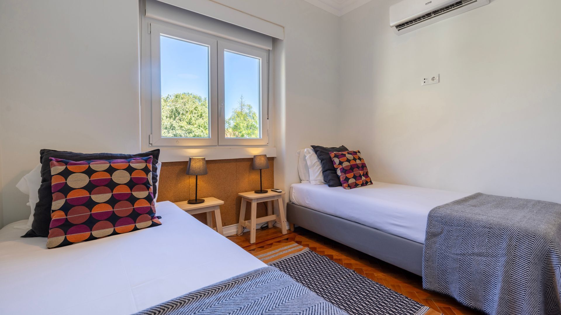 A small, well-lit room with two beds, a window, a heater, and a rug.
