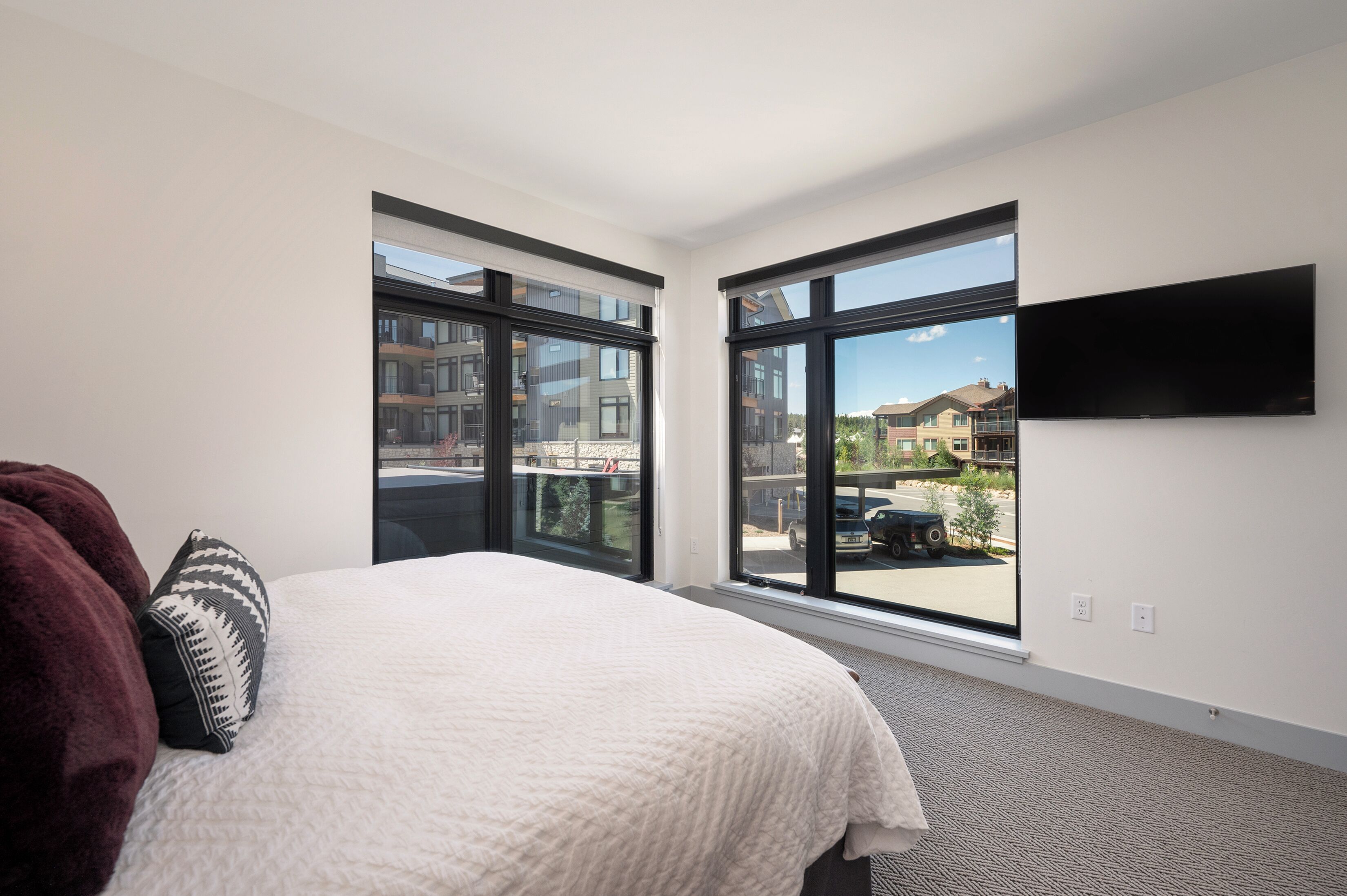 A modern hotel room with a large bed, a flat-screen TV mounted on the wall, and large windows offering a view of the outside.