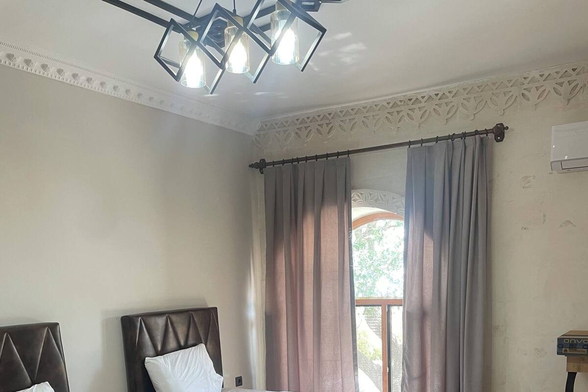 A room with two beds, a window with curtains, a ceiling light fixture, and a small table.