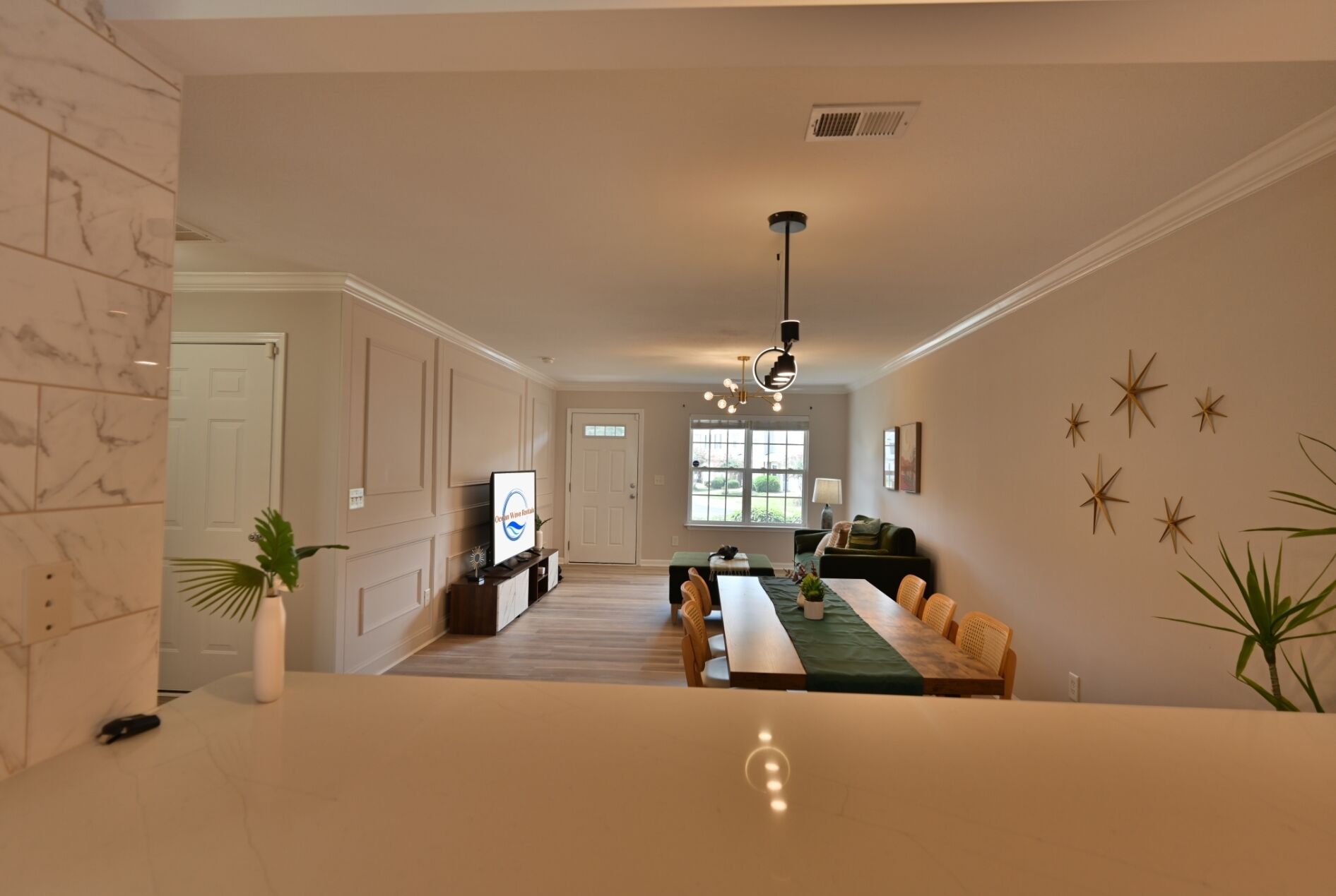 A spacious living room with a large window, a dining area with a green table, and a kitchen area with a TV.