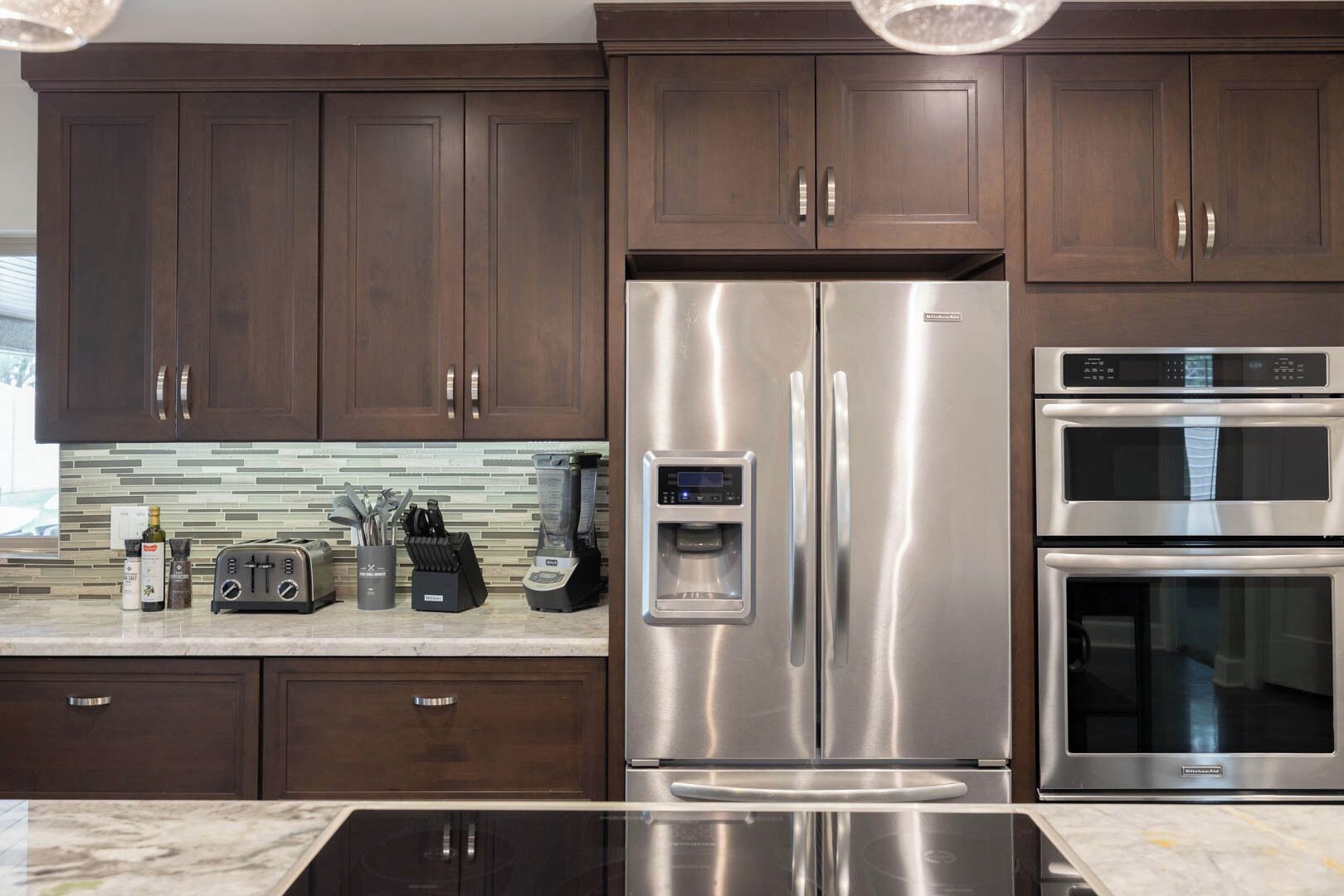 A modern kitchen with dark wooden cabinets, stainless steel appliances including a refrigerator and oven, a built-in coffee maker, and a toaster.