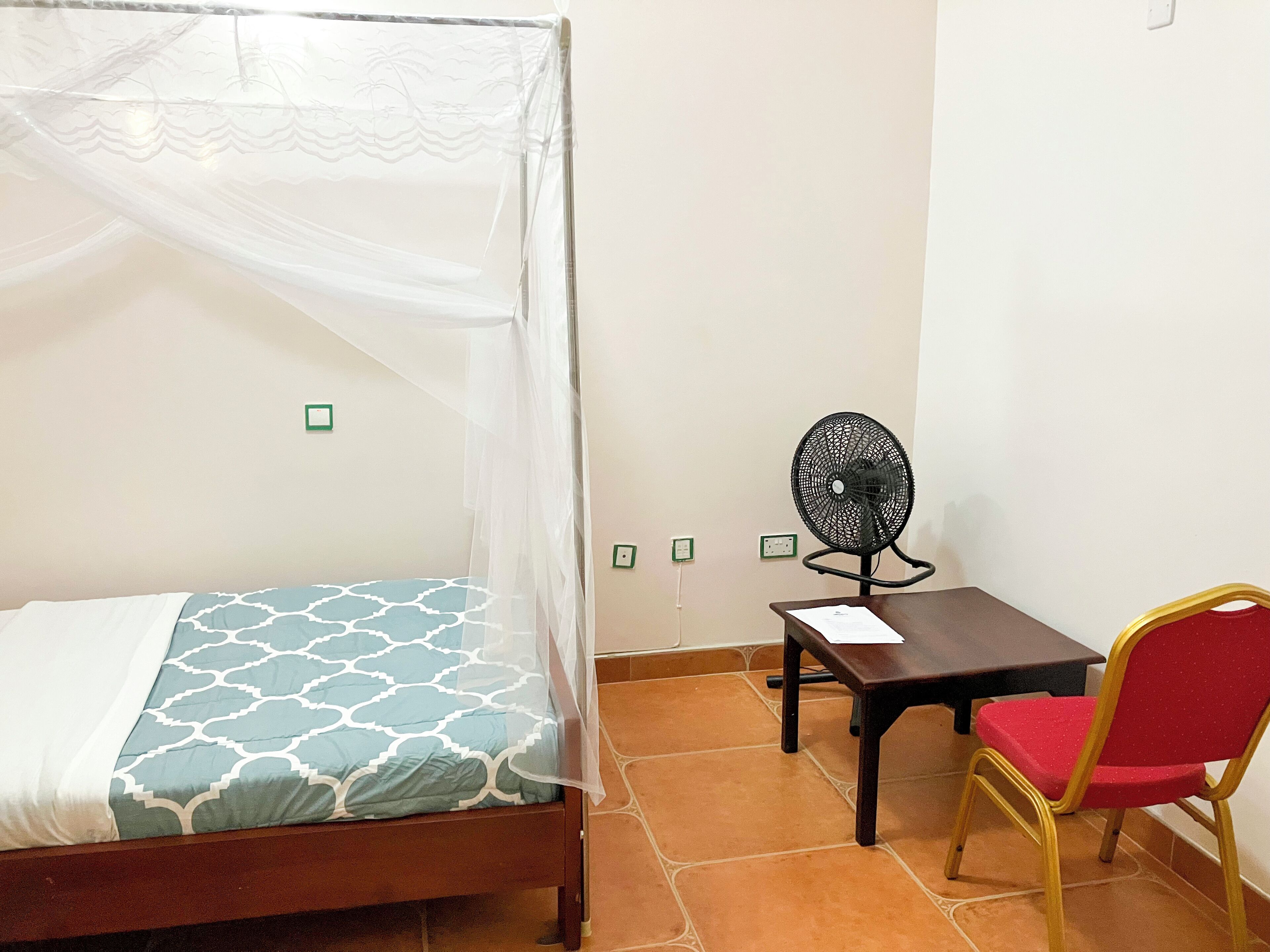 A bedroom with a bed, a desk, a chair, and a standing fan.