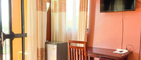 A room with a wooden desk, a chair, a television mounted on the wall, and a sliding glass door with curtains.