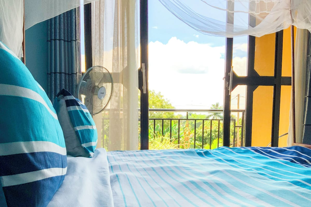 A bed with striped bedding, a standing fan, and a view of greenery through an open balcony door.