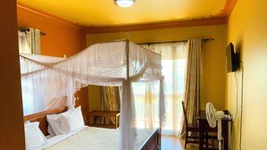 A room with a bed, a bedside table, a chair, a standing fan, and a television on the wall. - Hendo Hotel (Entebbe)