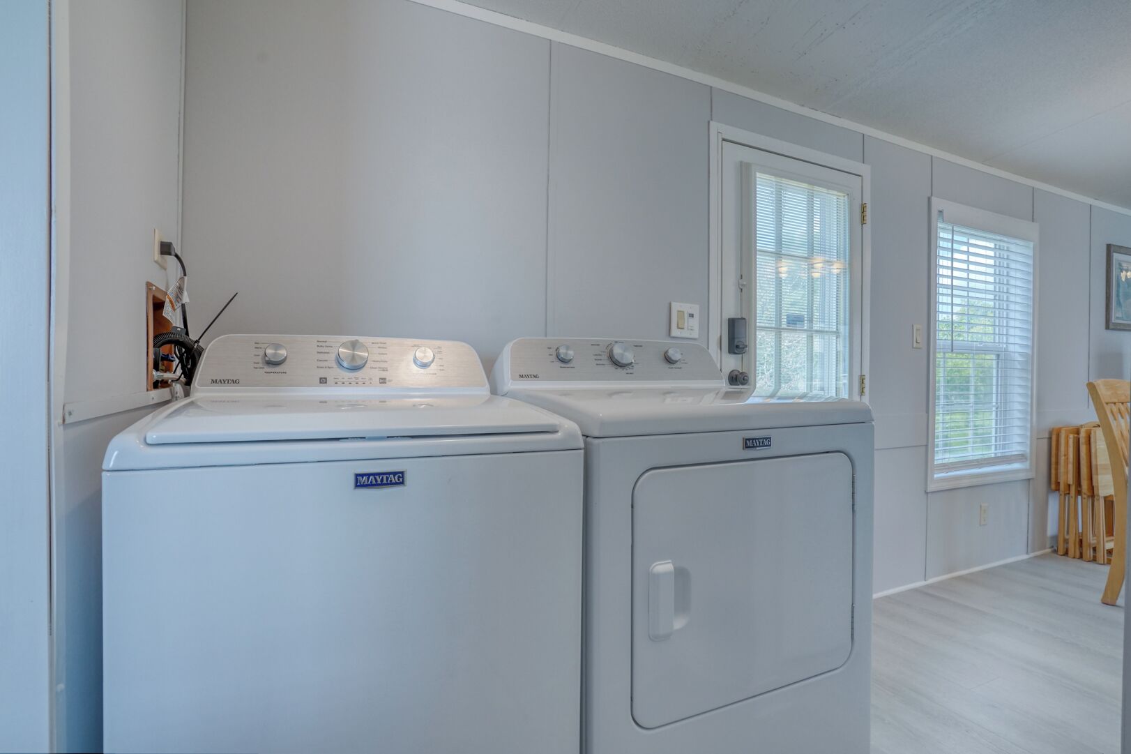 Une buanderie équipée de deux machines à laver et deux sèche-linge Maytag, d’une chaise en bois et d’une fenêtre avec des stores.