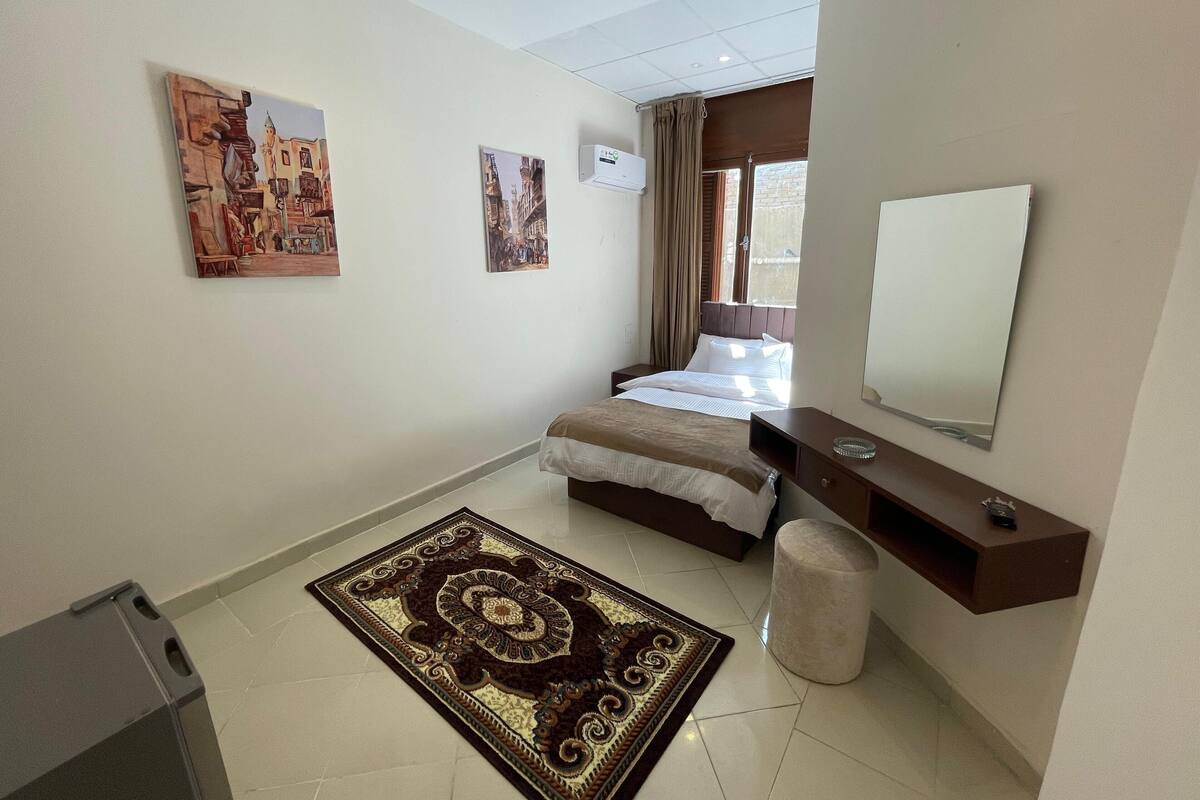 A hotel room with a bed, a bedside table, a mirror, a window with curtains, and a wall-mounted air conditioner.