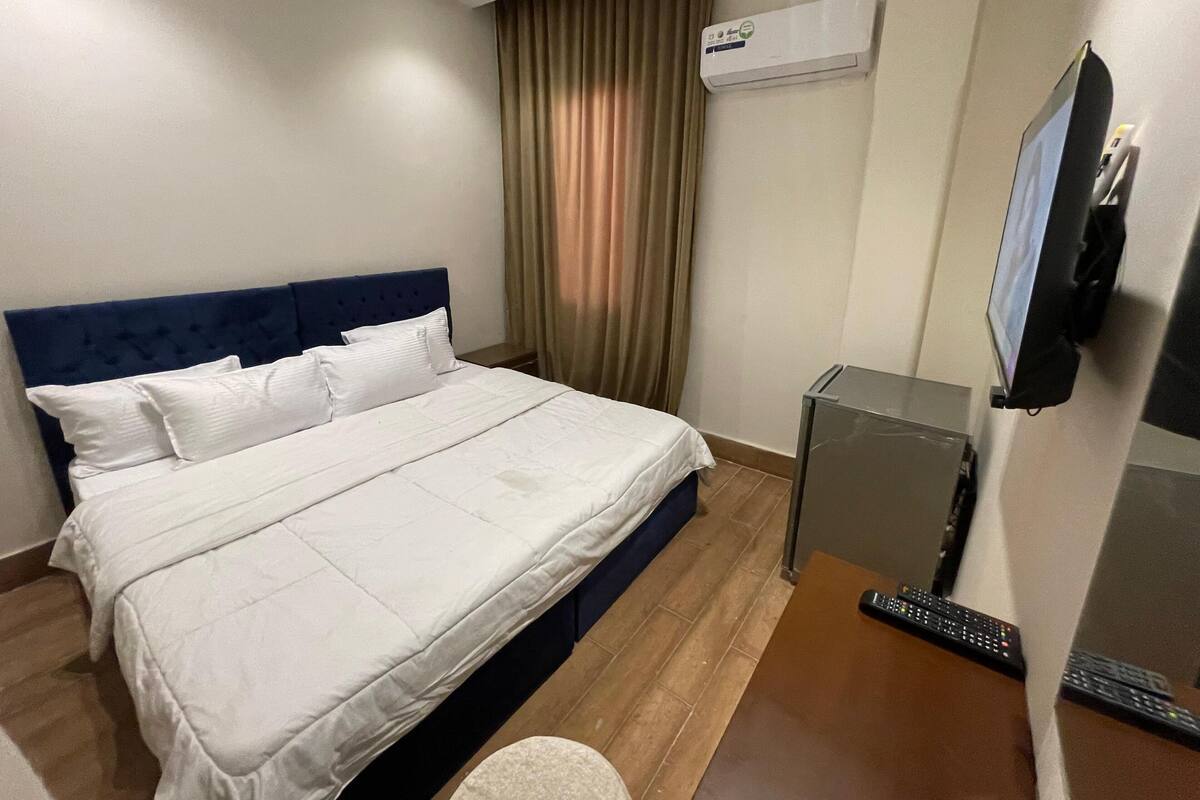 A hotel room with a bed, a TV mounted on the wall, and an air conditioning unit.