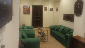 Una sala de estar con dos sofás verdes, una mesa de centro de cristal y un mueble de madera oscura. Se observan obras de arte colgadas en la pared y una puerta al fondo.