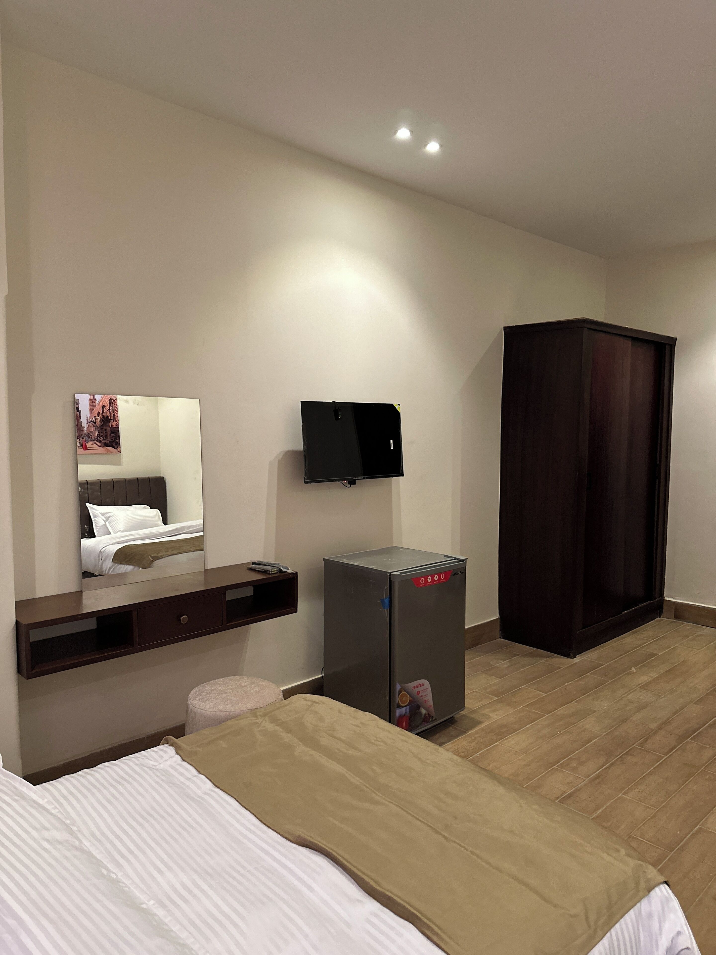 A hotel room with a bed, a small refrigerator, a dark wardrobe, a flat-screen TV mounted on the wall, and a mirror reflecting the room.
