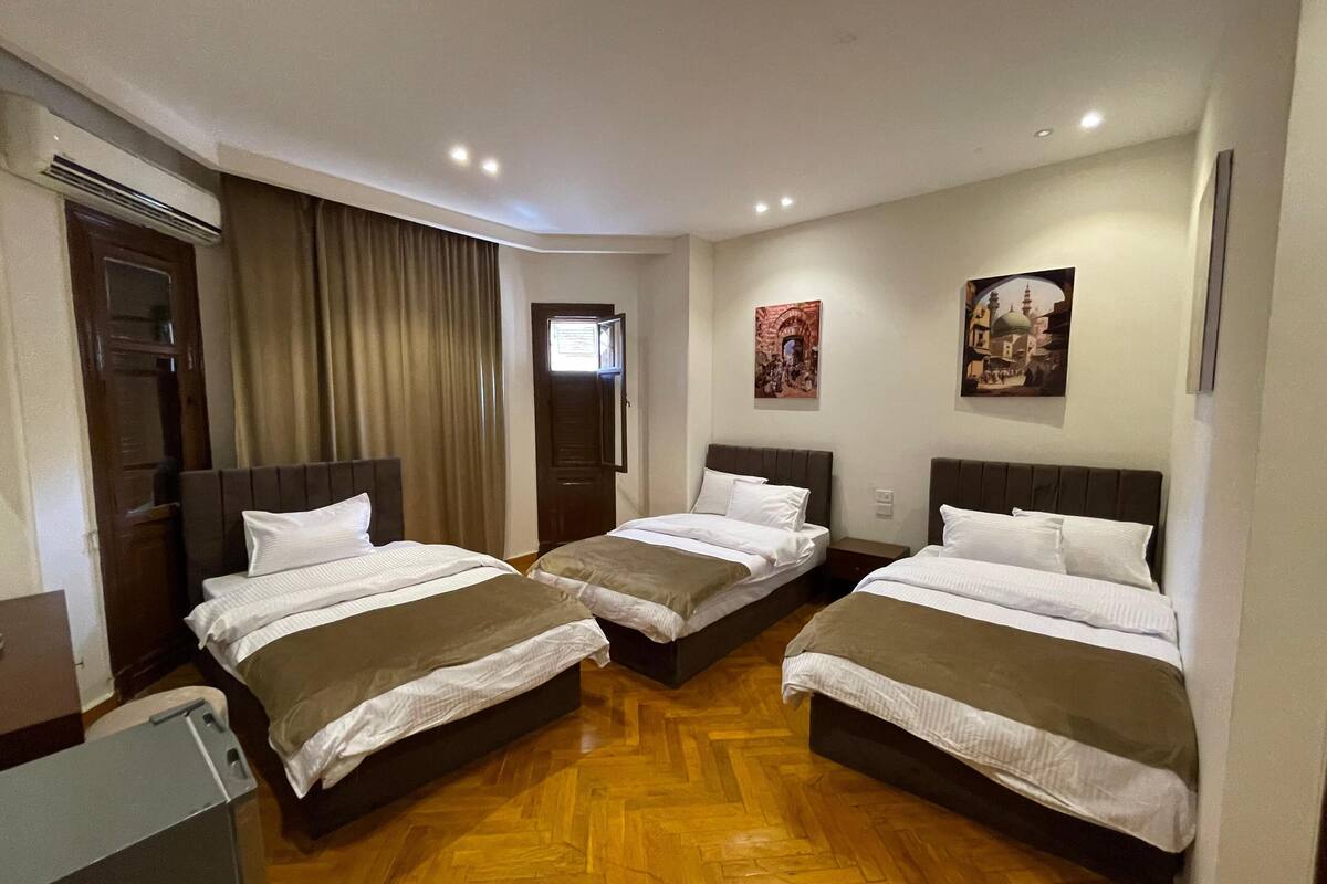 A hotel room with three single beds, wooden flooring, a door with curtains, and two wall-mounted pictures.