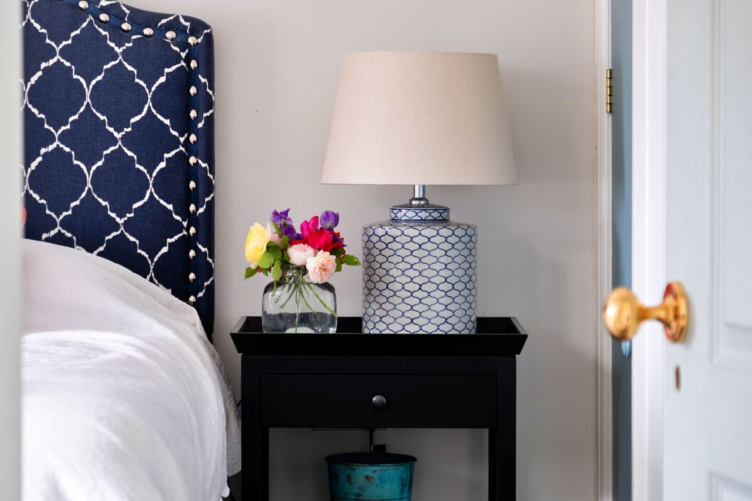 A neatly made bed with a white comforter, a dark blue patterned pillow, a bedside table with a lamp, a vase with flowers, and a decorative metal container.