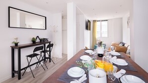 Un espace repas moderne avec une longue table dressée pour quatre personnes, une petite table d’appoint agrémentée d ’une plante et un téléviseur fixé au mur.