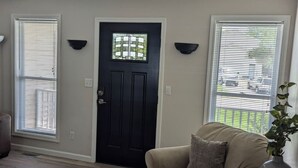 A living room with a dark door, two windows with blinds, a beige sofa, and a plant on a table.
