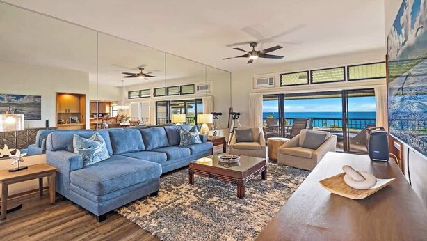 A spacious living room with a blue sofa, a wooden coffee table, and a view of the ocean through large windows. - Panoramic Ocean View Kapalua Villa #321 (Lahaina)