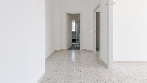 Un pasillo con paredes blancas y suelo de baldosas que conduce a un baño con ducha e inodoro visibles a través de una puerta.