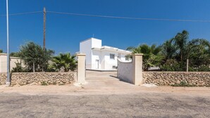 Un edificio blanco con entrada de pared de piedra, rodeado de palmeras y un cielo azul despejado.