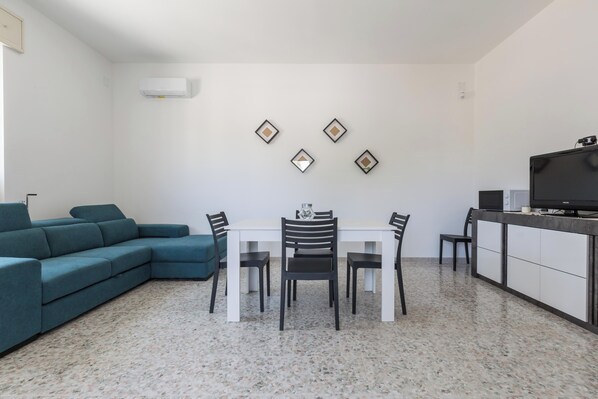 A modern living room with a sectional sofa, a dining table with chairs, a TV on a cabinet, and decorative wall art.