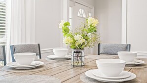 Una mesa de comedor con platos y tazones blancos, un florero con flores blancas y sillas con cojines grises.