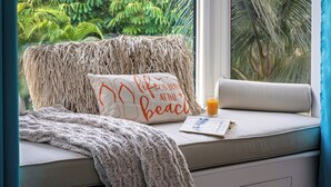 Eine gemütliche Leseecke mit einem gewobenen Pouf, einem Kissen mit Strandmotiv und einem Glas Orangensaft auf einer Fensterbank.