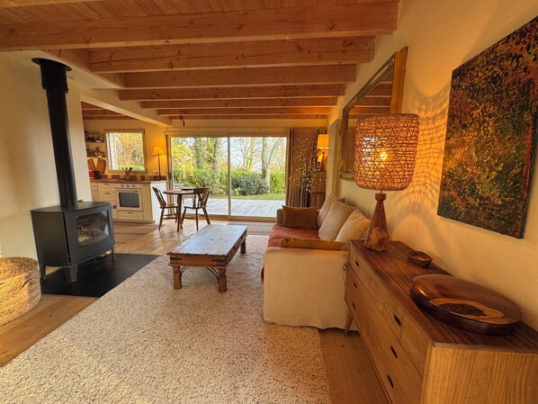 A cozy living room with a fireplace, wooden furniture, a dining area, and a large window offering a view of the outdoors.