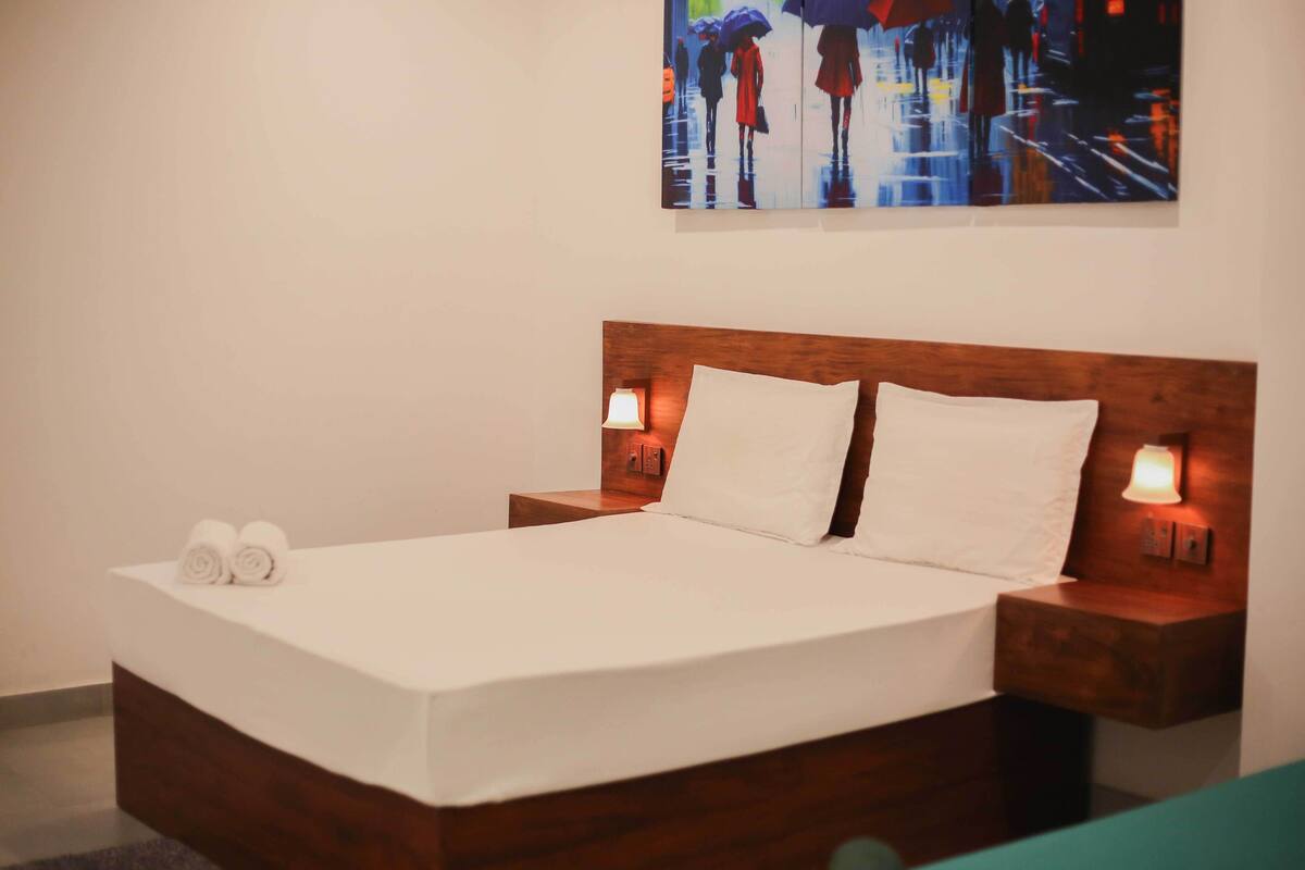 A neatly made bed with white linens, two bedside lamps, and a painting of people with umbrellas on the wall.