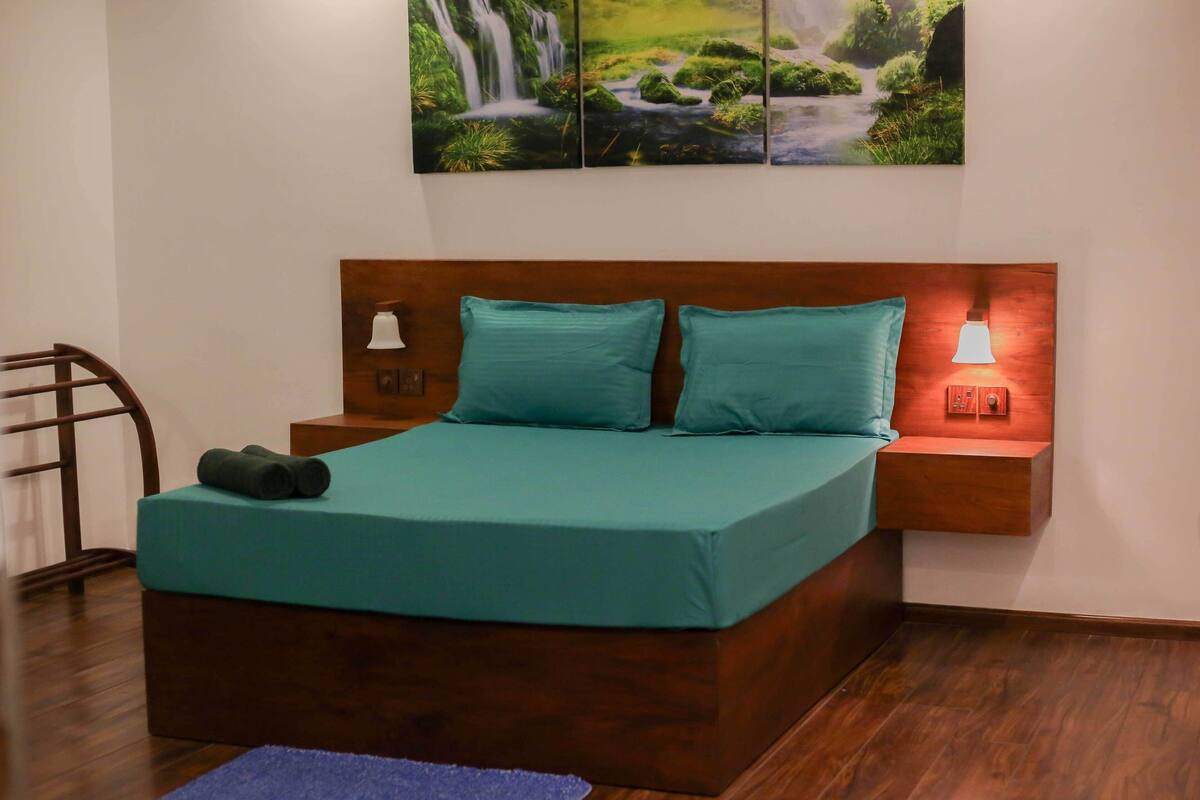 A neatly made bed with a teal bedspread and pillows, wooden headboard, two bedside tables with lamps, and a wall-mounted painting of a waterfall.