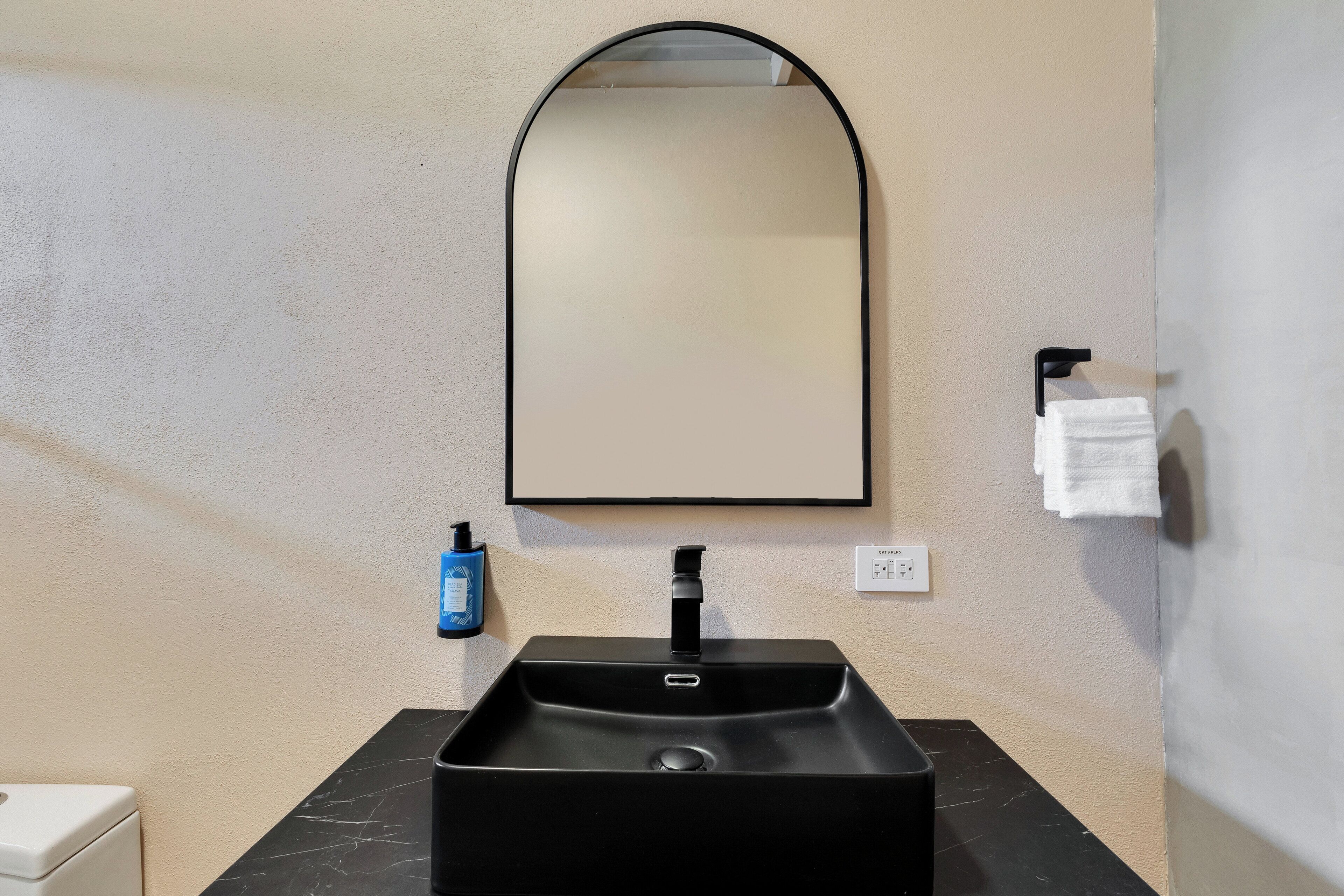 A bathroom with a black sink, a mirror above it, a towel hanging on a hook, and a bottle of hand soap on the wall.