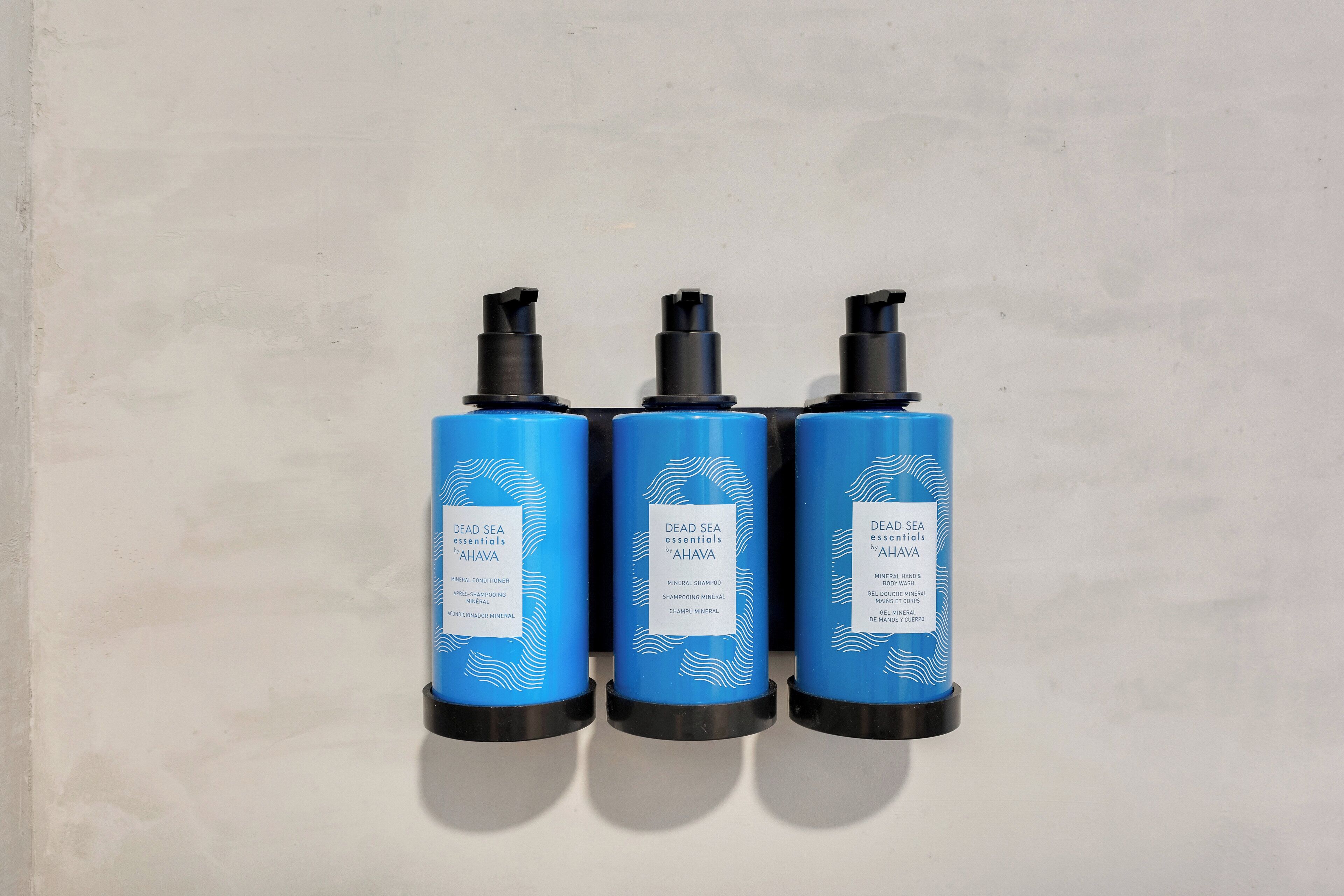 Three blue bottles with black caps labeled 'Dead Sea Essentials' against a plain wall.