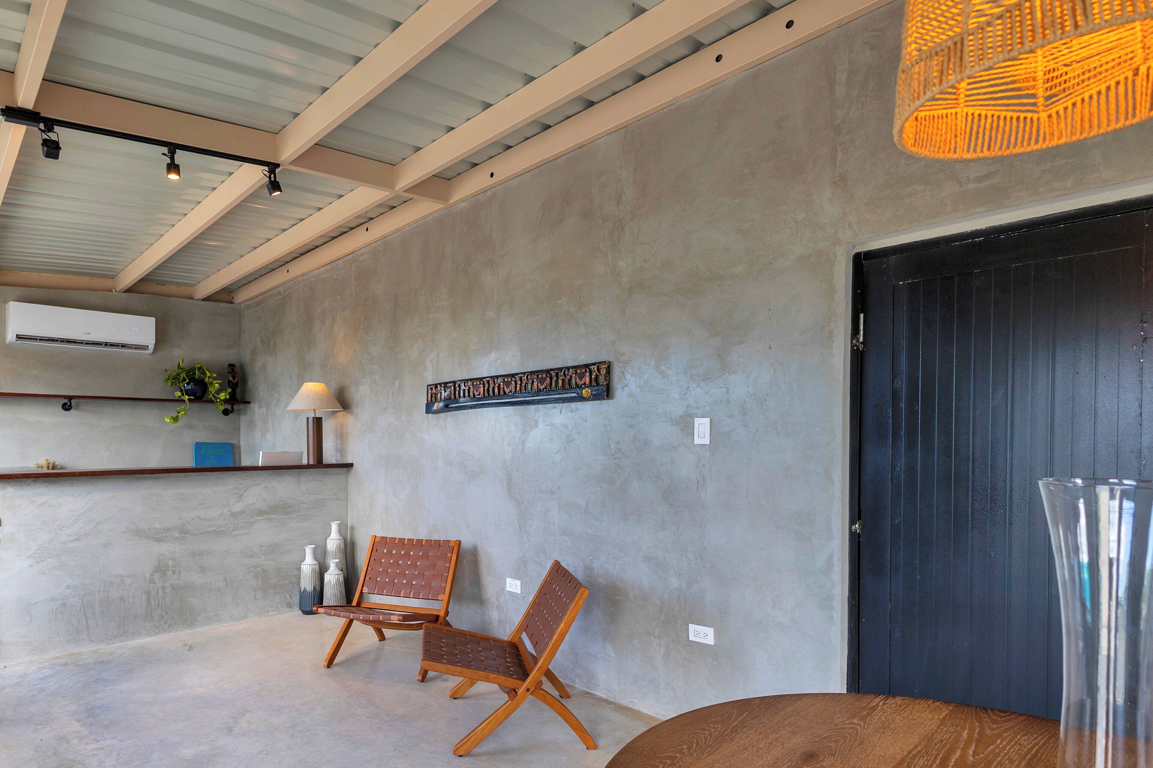 A room with a concrete wall, a wooden chair, a table, a hanging light fixture, and a door.