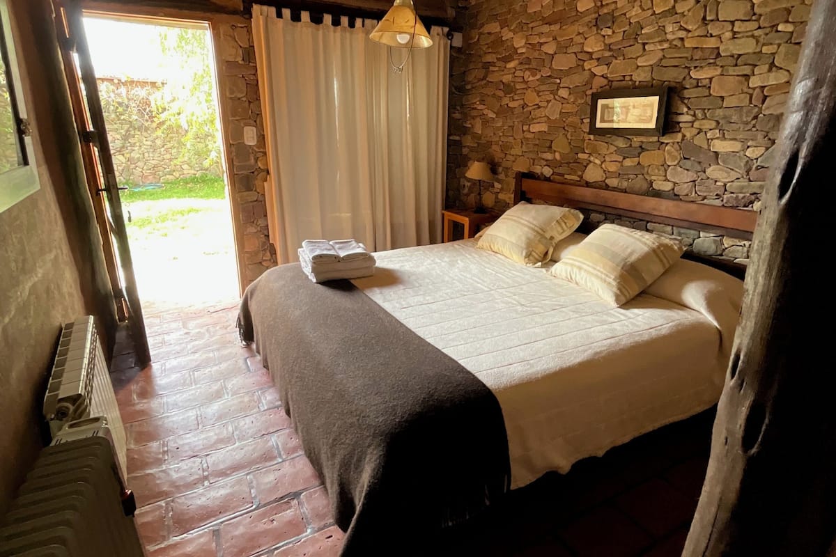 Un dormitorio con pared de piedra, una cama con colcha blanca, una lámpara colgante y un ventanal con cortinas.
