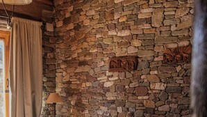 A bedroom with a stone wall, a bed with a red blanket, a lamp on a wooden nightstand, and a window with curtains. - HOSTERIA REFUGIO DEL PINTOR (Tilcara)