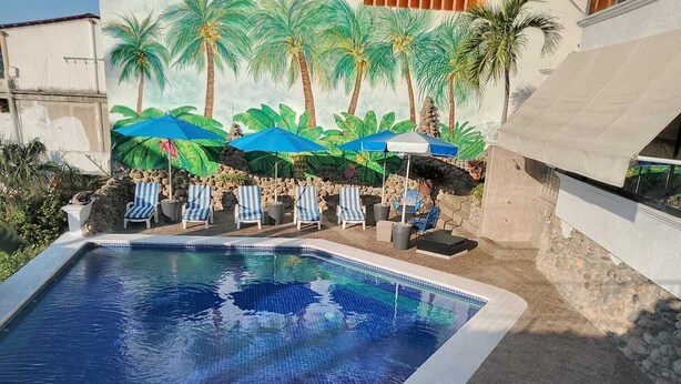 Una piscina con borde de azulejos, rodeada de sillas de descanso y sombrillas, con un mural de palmeras al fondo.