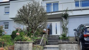 Ein zweistöckiges Gebäude mit einer Treppe zum Eingang, umgeben von einem Garten mit blühenden Pflanzen und einem Baum.