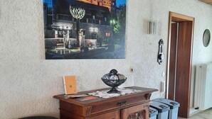 Ein Wohnzimmer mit einem dunkelblauen Sofa, einer Holzkassette und einem Gemälde eines Gebäudes. Im Hintergrund befinden sich zwei Mülleimer und eine Tür.