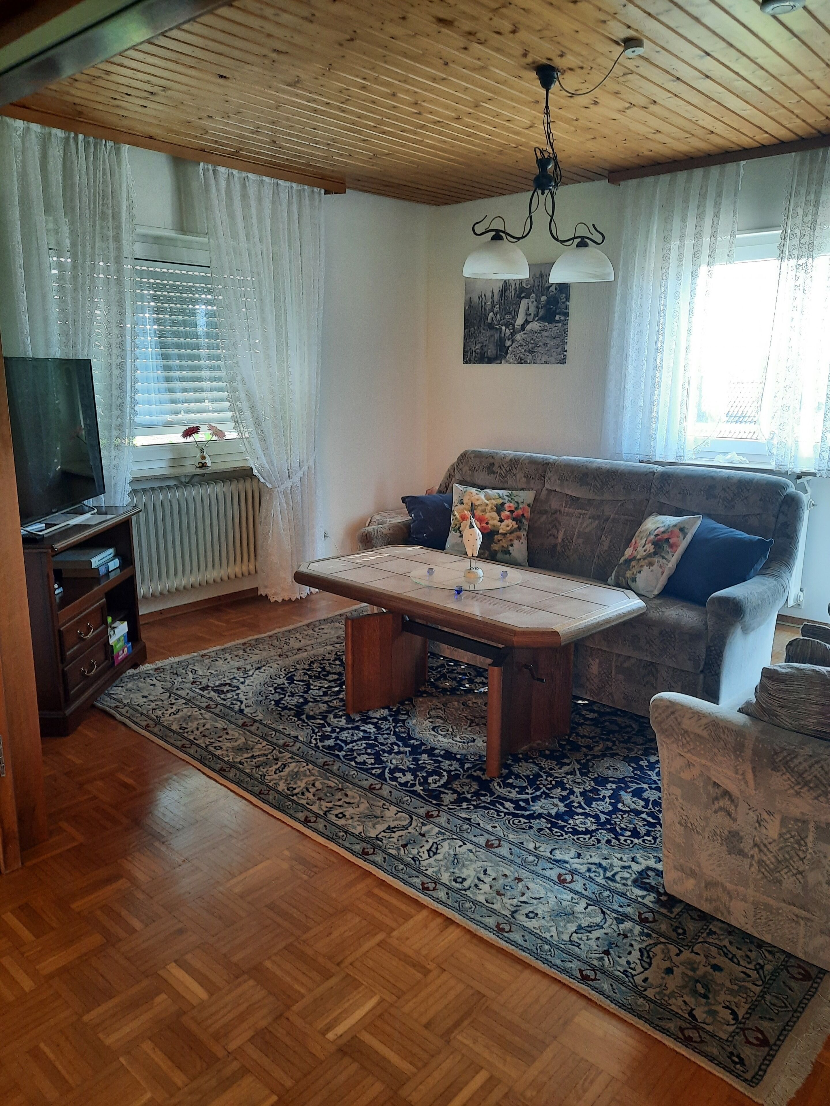Een woonkamer met een houten plafond, een bank, een salontafel, een televisie en een kamerkleed met dessin.