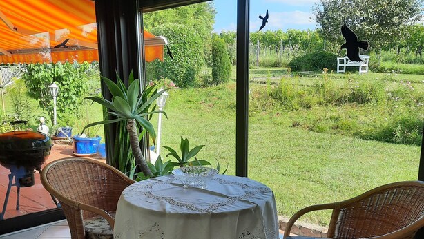 Ein Patio mit Korbstühlen und einem Tisch, bedeckt mit einer Spitzenleinentischdecke, mit Blick auf einen Garten mit einer weißen Bank und Vogelskulpturen.