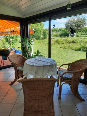 A patio area with wicker chairs and a table covered with a lace tablecloth, overlooking a garden with a white bench and bird sculptures.