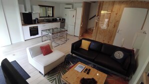 Un salón moderno con un sofá negro, un sillón blanco y una mesa de centro de madera. Se ve en el fondo una cocina con armarios blancos y un comedor con una mesa y sillas de cristal.