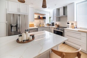 Una cocina moderna con armarios blancos, una isla de mármol, electrodomésticos de acero inoxidable y una silla de madera.