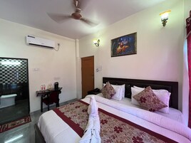 A hotel room with a bed, a desk, a chair, a mirror, an air conditioner, a door, and a wall-mounted light fixture.