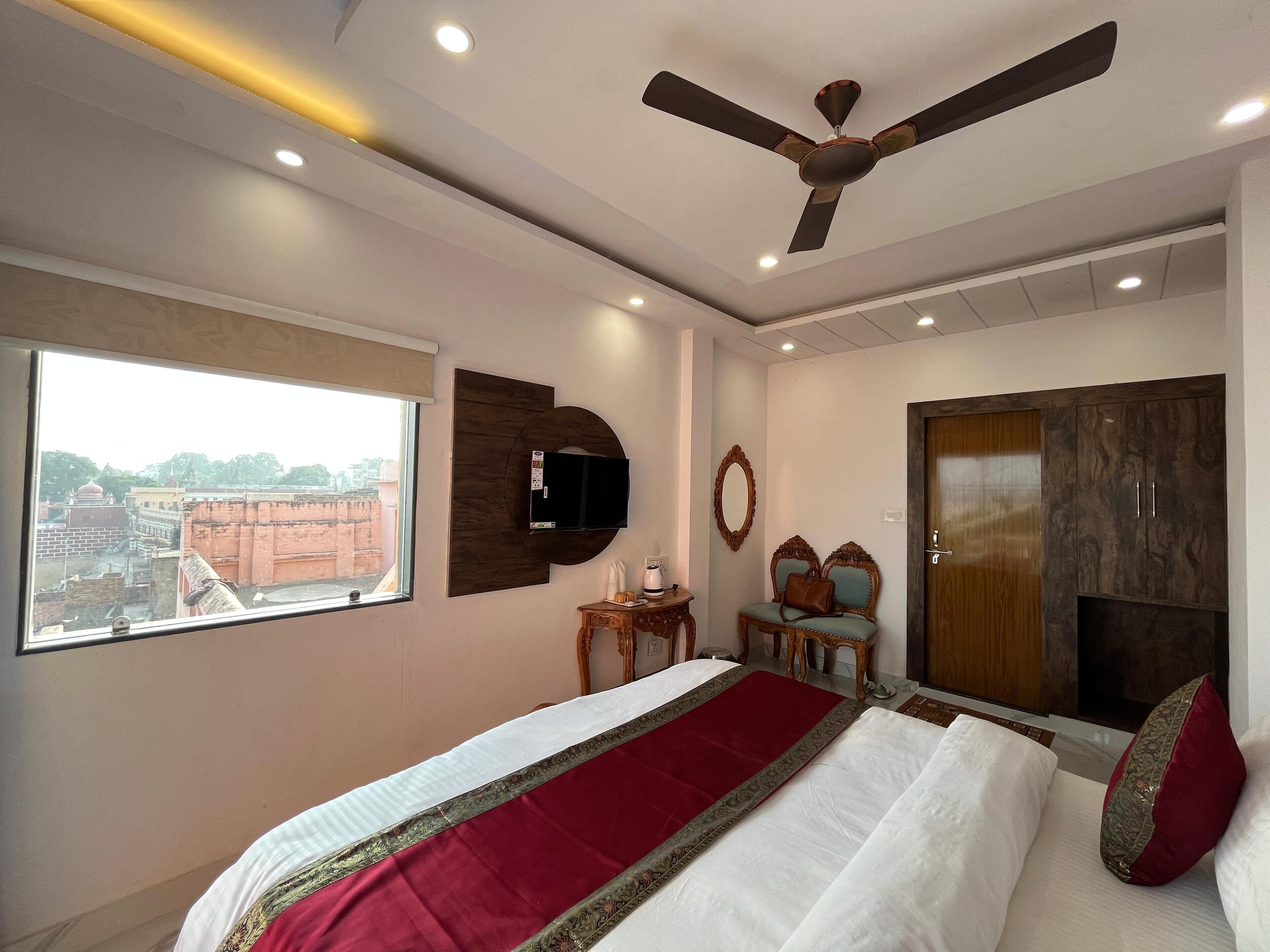Un dormitorio con cama, ventilador de techo, televisor montado en la pared, ventana con vistas a edificios y armario.