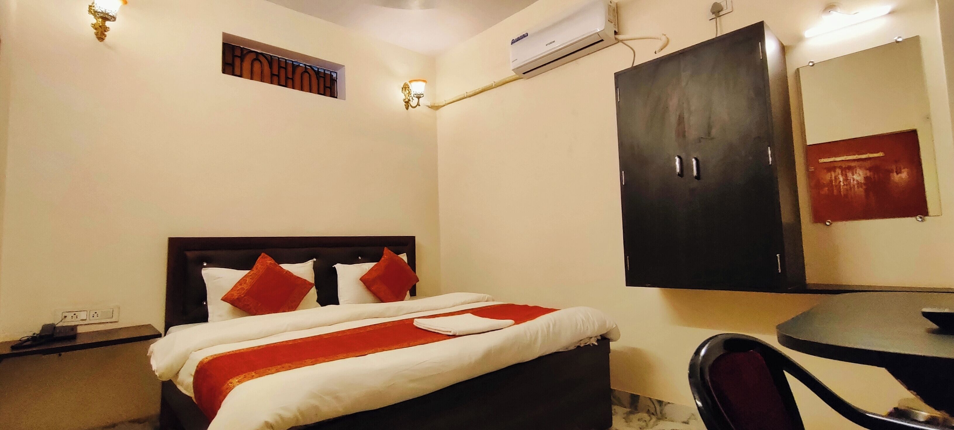 A hotel room with a bed, a desk, a chair, a wardrobe, and an air conditioning unit.