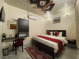 A bedroom with a bed, a desk, a chair, a ceiling fan, an air conditioner, a trash bin, a rug, and a painting on the wall.